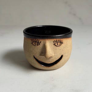 May include: A small, tan ceramic cup with a smiling face design. The cup features a black rim and a black interior. The face has detailed eyes, a nose, and a curved smile. The cup is likely handmade.