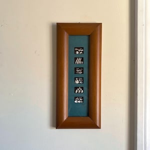 May include: A framed artwork featuring six small, rectangular compositions. Each composition contains a unique arrangement of small, white objects against a teal background. The frame is made of wood with a warm, brown finish. The artwork is mounted on a white wall.