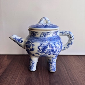 May include: A blue and white ceramic teapot with a lid, spout, and handle. The teapot is decorated with floral and fish motifs. It has three small legs and a loop on the lid. The teapot is on a dark wooden surface.