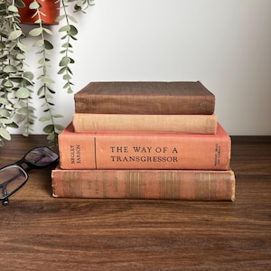 May include: A stack of four vintage books with aged covers in shades of brown and peach. The book titled "The Way of a Transgressor" is visible. A pair of black-framed glasses rests on the wooden surface.