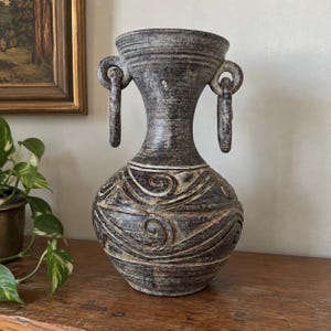 May include: A dark gray ceramic vase with a textured, mottled finish and a wide, flared rim. The vessel features two prominent ring-shaped handles attached to the neck and a body decorated with raised, swirling patterns that wrap around the base.