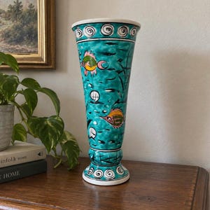 May include: A tall, flared ceramic vase featuring a vibrant turquoise glaze with hand-painted fish motifs in yellow, pink, and green. The design includes black line details, circular patterns at the rim and base, and a wide, open top opening.