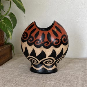 May include: A rounded ceramic vase featuring a wide, notched top opening. The exterior displays a hand-painted pattern of black swirls and triangular shapes against a cream-colored background, with a band of terracotta orange at the upper rim.