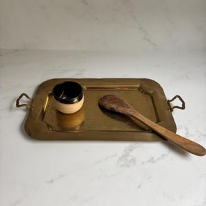 May include: A rectangular brass tray with handles, holding a small black and beige bowl and a wooden spoon. The tray has a decorative edge and a polished finish. The spoon is made of dark wood.