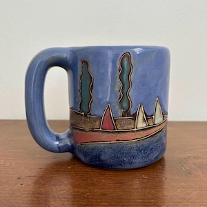 Puede incluir: Una taza de cerámica con esmalte azul que presenta un diseño de paisaje. El exterior muestra dos árboles altos de color verde oscuro y varias formas triangulares pequeñas en rojo, blanco y azul, delineadas en marrón oscuro sobre un fondo azul texturizado.