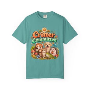 Op de afbeelding: Tealkleurig T-shirt met de tekst "Critter Committee" en een afbeelding van een rode panda, kat, hond, egel en kikker. Het ontwerp bevat bloemen, paddenstoelen en een vlinder. Het shirt is gemaakt van een zacht materiaal.
