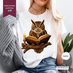 Può includere: Una t-shirt bianca con una stampa grafica di un gufo marrone con occhiali, appollaiato su un ramo di un albero mentre legge un libro aperto accanto a una candela accesa. Il gufo ha piume dettagliate e occhi gialli, e il libro ha pagine visibili.