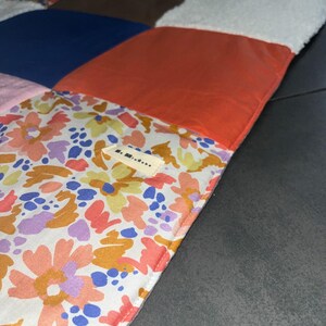 Puede incluir: Una colcha de patchwork con diversos paneles de tela cuadrados en naranja sólido, azul marino y material blanco texturizado, junto a tela con estampado floral de flores rosas, amarillas y azules. Una pequeña etiqueta rectangular dice S. Mistene.