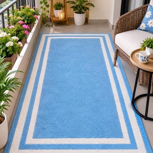 May include: A rectangular area rug featuring a solid light blue center framed by a thick white border and an outer light blue border. The rug has a textured, soft-looking pile and is positioned on a flat floor surface in an outdoor space.