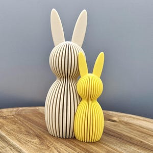 Op de afbeelding: Twee decoratieve houten konijnenbeeldjes. Eén is gebroken wit, de andere is geel. Beide hebben een gleufontwerp en lange oren. Ze staan op een houten oppervlak tegen een grijze achtergrond. De konijnen zijn een charmant paas- of lentedecoratie-item.
