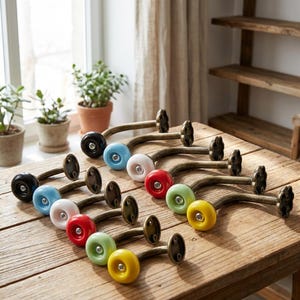 Puede incluir: Una colección de ganchos de pared decorativos con una estética vintage. Cada gancho presenta un brazo de metal curvado en tono bronce y un pomo de cerámica de colores en negro, azul, blanco, rojo, verde y amarillo. Los ganchos están dispuestos sobre una superficie de madera.