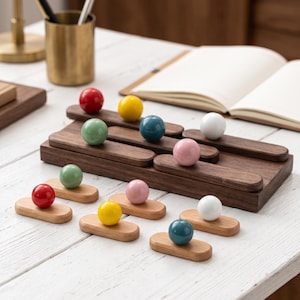 May include: A set of wooden organizers featuring smooth, dark walnut rectangular bases and smaller light wood oval bases, each topped with a single colorful sphere in red, yellow, sage green, teal, white, or soft pink, arranged on a white surface.