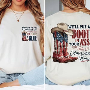 May include: An off-white t-shirt featuring a small graphic on the front chest with a cowboy boot and the text "BROUGHT TO YOU COURTESY OF THE RED, WHITE & BLUE". The back displays a large graphic of a cowboy hat and boots with the text "WE'LL PUT A BOOT IN YOUR ASS The American Way".