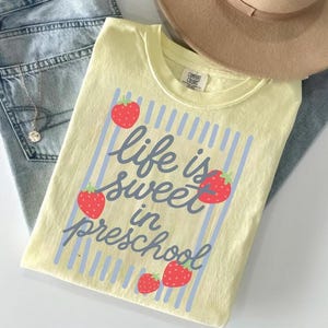 May include: A pale yellow short-sleeve t-shirt featuring a graphic of blue vertical stripes and four red strawberries. The text "life is sweet in preschool" is printed in a dark blue script font across the center of the shirt design.