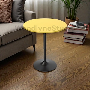 May include: A modern side table with a round, yellow tabletop and a black, pedestal base. The table is set on a hardwood floor, next to a sofa and a stack of books. The table's design is simple and contemporary.