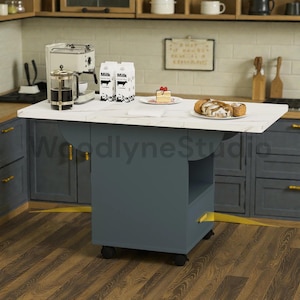 May include: A gray kitchen island with a white marble-look countertop. The island has a lower shelf and is on wheels. Items on the countertop include a coffee maker, milk cartons, pastries, and a cake.