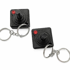 Puede incluir: Dos llaveros cuadrados negros con forma de joysticks de videojuegos retro. Cada llavero cuenta con un mando central negro, un botón rojo circular en la esquina superior izquierda y una anilla de metal plateado unida por una cadena corta.