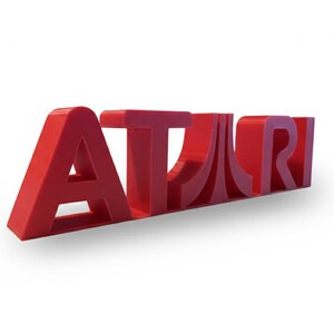May include: A red, three-dimensional "ATARI" logo, with the letters in a bold, stylized font. The logo is angled slightly, casting a shadow on the white background. This item is a decorative piece.