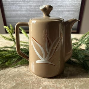 May include: A tan ceramic coffee pot with a lid and a handle. The pot has a white and brown floral design on the front. The spout is on the right. The lid has a small, rounded knob. The pot is on a marble surface.