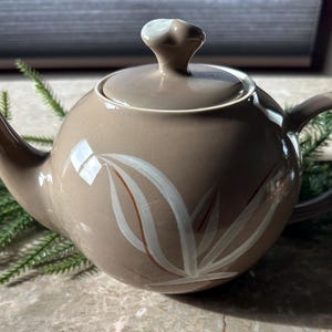 May include: A taupe ceramic teapot with a matching lid and handle. The teapot features a white and brown floral design on the side. The spout and handle are curved. The lid has a small, rounded knob. The teapot is on a marble surface.