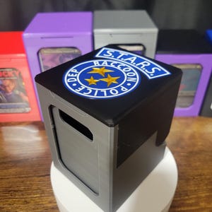 May include: A gray and black plastic deck box featuring a circular S.T.A.R.S. Raccoon Police Department emblem on the top lid. The emblem includes three gold stars on a blue background with white text. The box has a rectangular cutout on the front.