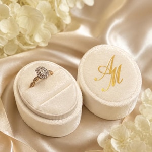 May include: An oval cream-coloured velvet ring box with a gold monogram AM on the lid, containing a gold ring with a central oval stone surrounded by a halo of smaller stones. The box has a single slot for the ring and a matching lid.