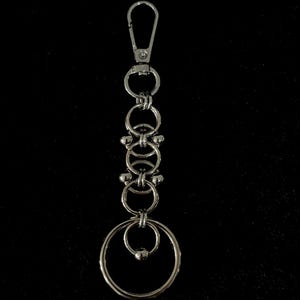 Könnte beinhalten: Ein silberfarbener Schlüsselanhänger mit einem Karabiner oben und mehreren miteinander verbundenen Ringen. Das Design umfasst eine Reihe kleiner dekorativer Elemente und einen größeren, offenen Ring unten. Der Hintergrund ist einfarbig schwarz.
