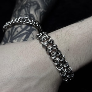 May include: A silver chainmail bracelet with a woven design. The bracelet is made of small, interlocking metal rings. The bracelet is shown on a wrist with a dark background. Another bracelet is visible in the background.