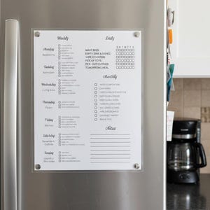 May include: A clear acrylic weekly and daily chore chart is attached to a stainless steel refrigerator door. The chart includes sections for daily, weekly, and monthly tasks, with check boxes for completion. The text includes "Weekly", "Daily", and "Monthly" headings.