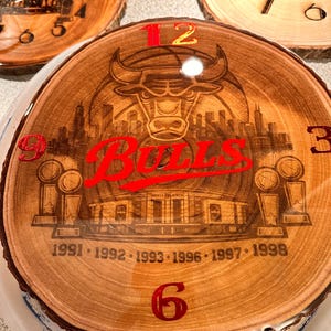 May include: A wooden clock featuring a Chicago Bulls design. The clock face is a cross-section of a tree trunk, with the Bulls logo, the word "Bulls", and the years of their championships. Red numerals mark the hours.