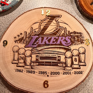 May include: A wooden clock featuring the Los Angeles Lakers logo, the Crypto.com Arena, and championship years. The clock has gold-colored numbers and a natural wood grain finish. The Lakers logo is in purple and brown.