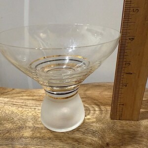May include: A clear cocktail glass with a frosted base and a flared bowl. The glass is adorned with gold and black bands. A wooden ruler is next to the glass.