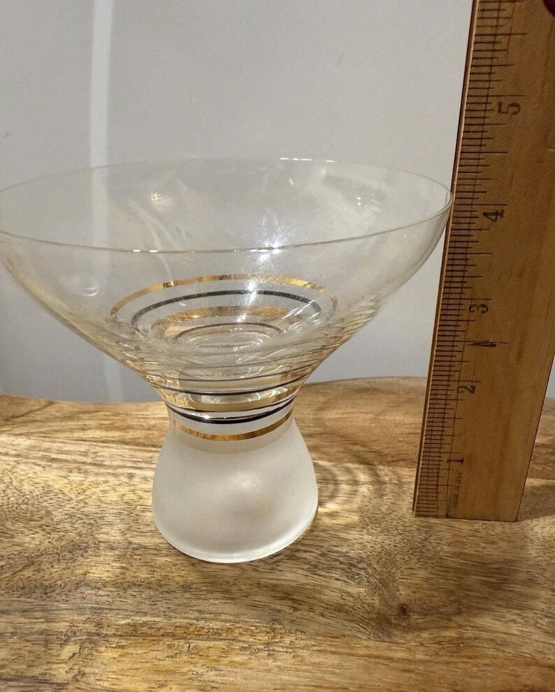 May include: A clear cocktail glass with a frosted base and a flared bowl. The glass is adorned with gold and black bands. A wooden ruler is next to the glass.