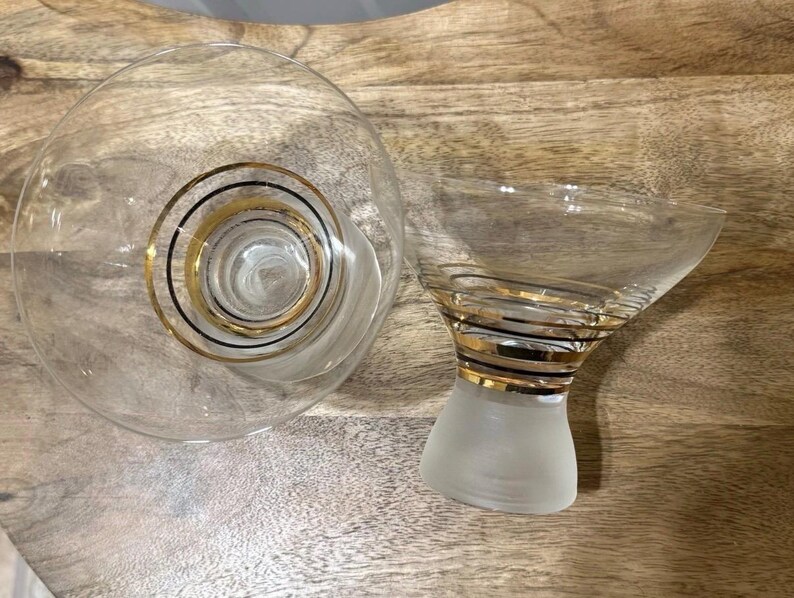 May include: Two clear glass martini glasses with gold and black spiral detailing. One glass is a wide, shallow bowl, and the other has a conical bowl atop a frosted base. The glasses are designed for cocktails.