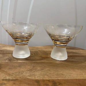 May include: Two clear martini glasses with gold and black spiral detailing around the stem and a frosted base. The glasses are on a wooden surface. The glasses have a classic, elegant design.