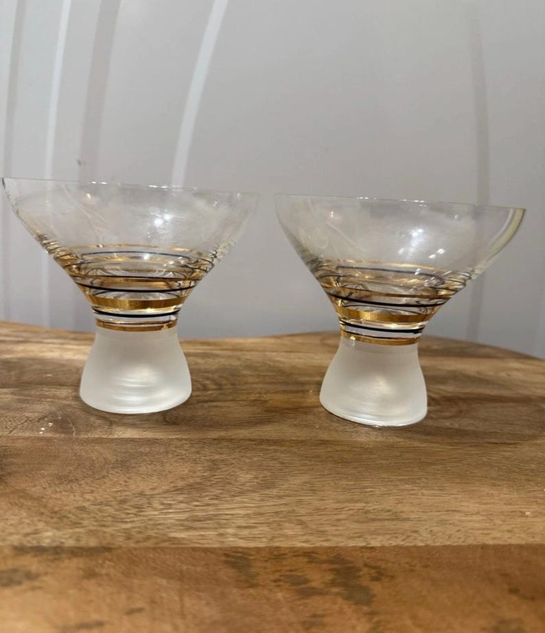 May include: Two clear martini glasses with gold and black spiral detailing around the stem and a frosted base. The glasses are on a wooden surface. The glasses have a classic, elegant design.