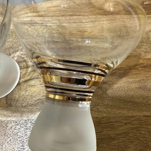 May include: Clear martini glass with a frosted base and a wide bowl. The glass is adorned with gold and black horizontal stripes around the stem. The glass is sitting on a wooden surface.