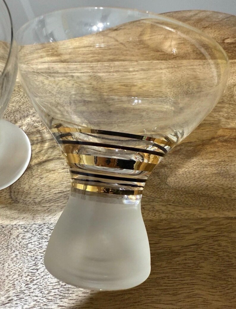 May include: Clear martini glass with a frosted base and a wide bowl. The glass is adorned with gold and black horizontal stripes around the stem. The glass is sitting on a wooden surface.