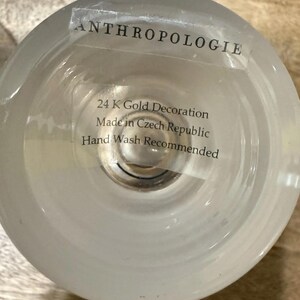 May include: A clear glass Anthropologie candle holder with a frosted finish. The label reads "24 K Gold Decoration Made in Czech Republic Hand Wash Recommended."