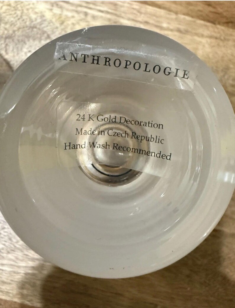 May include: A clear glass Anthropologie candle holder with a frosted finish. The label reads "24 K Gold Decoration Made in Czech Republic Hand Wash Recommended."