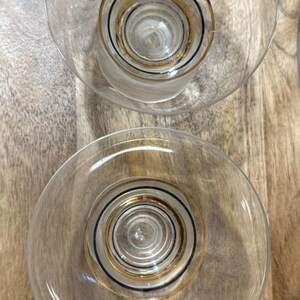 May include: Two clear glass cocktail glasses with a gold and black concentric circle design. The glasses have a wide, shallow bowl and a short stem. The glasses are on a wooden surface.