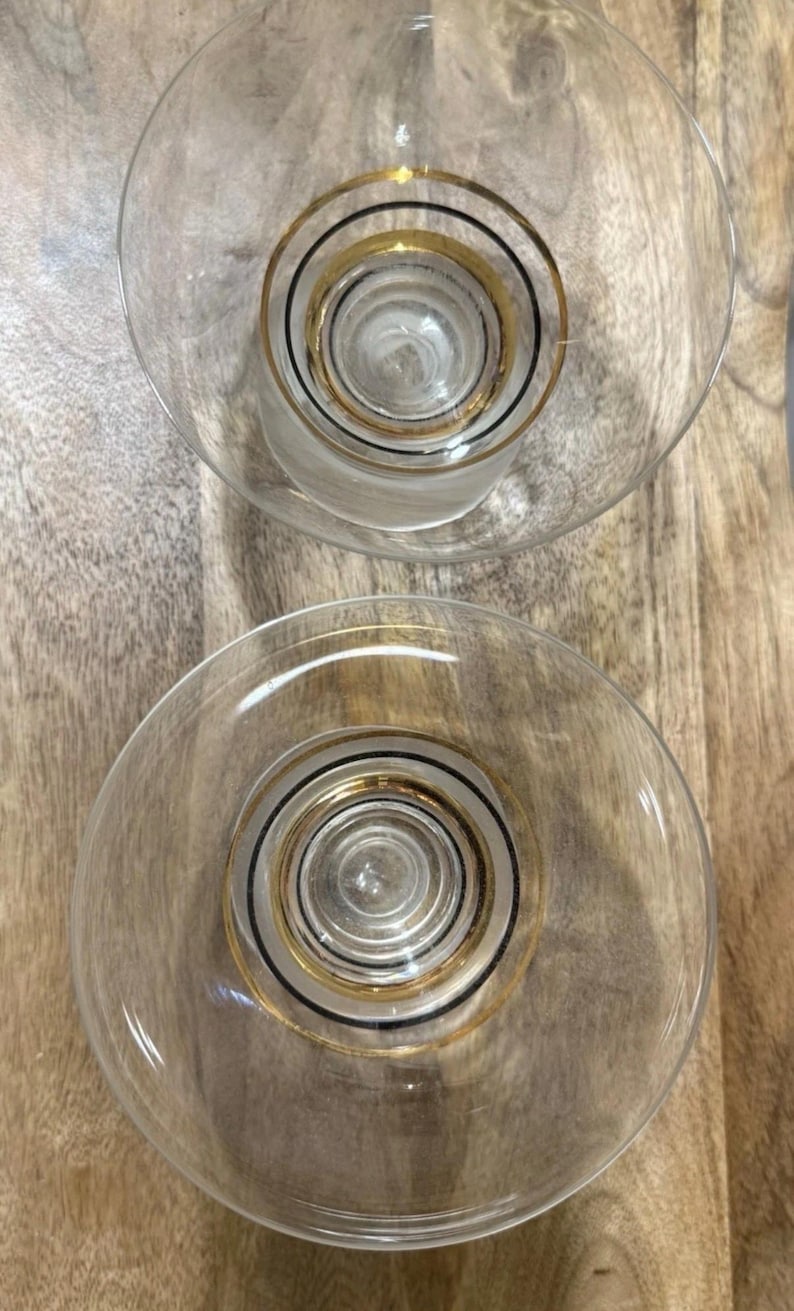 May include: Two clear glass cocktail glasses with a gold and black concentric circle design. The glasses have a wide, shallow bowl and a short stem. The glasses are on a wooden surface.