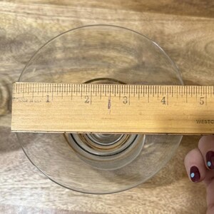 May include: A clear glass dish with a gold rim, measured with a wooden ruler. The ruler shows the dish is approximately 5 inches in diameter. The dish has a decorative pattern in the center.