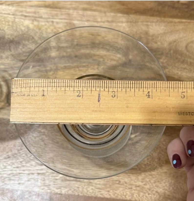 May include: A clear glass dish with a gold rim, measured with a wooden ruler. The ruler shows the dish is approximately 5 inches in diameter. The dish has a decorative pattern in the center.