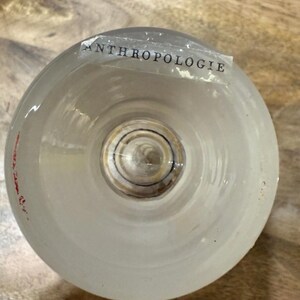 May include: A clear, frosted glass with the word "ANTHROPOLOGIE" printed on a label. The glass has a rounded shape and a circular base. The glass is sitting on a wooden surface.