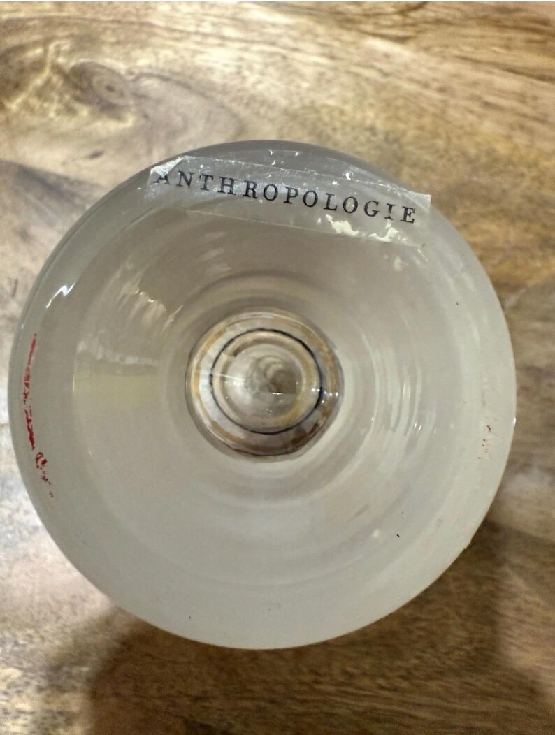 May include: A clear, frosted glass with the word "ANTHROPOLOGIE" printed on a label. The glass has a rounded shape and a circular base. The glass is sitting on a wooden surface.