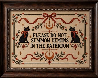 Könnte beinhalten: Ein gerahmtes Kreuzstichbild mit dem Text PLEASE DO NOT SUMMON DEMONS IN THE BATHROOM. Das Design zeigt zwei schwarze Katzen, orangefarbene Mondsicheln, rote Schleifen und florale Motive auf einem beigen Stoffhintergrund.