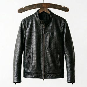 Könnte beinhalten: Schwarze Lederjacke mit strukturierter Krokodilmusterung. Die Jacke hat einen Stehkragen, einen Frontreißverschluss und mehrere Reißverschlusstaschen. Sie hängt an einem Holzbügel vor weißem Hintergrund.