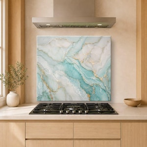 May include: A square kitchen splashback panel featuring a marbled pattern in shades of white, soft teal, and light green, accented with thin, flowing gold veins. The panel is mounted on a wall behind a black gas hob and a stainless steel extractor hood.