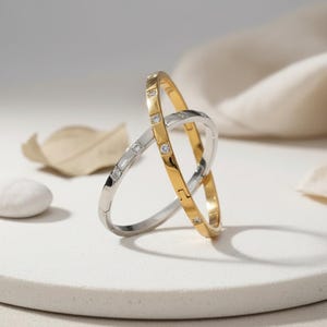 May include: Two intertwined bangles, one gold-toned and one silver-toned, each adorned with small, sparkling gemstones. The gold bangle is on the right, and the silver bangle is on the left. The bangles are on a white surface.